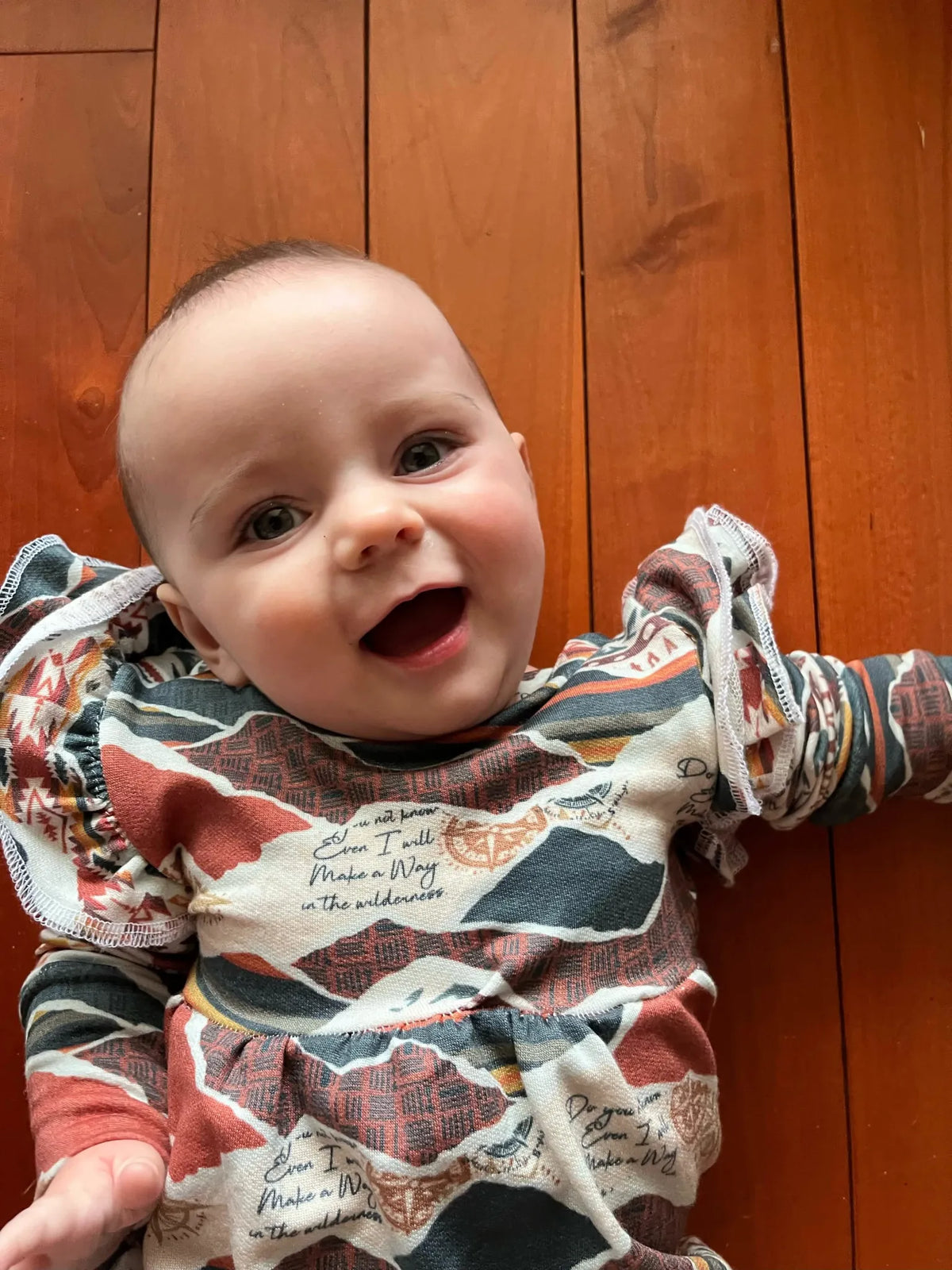 A baby in a colorful patterned outfit with text on a wooden floor.