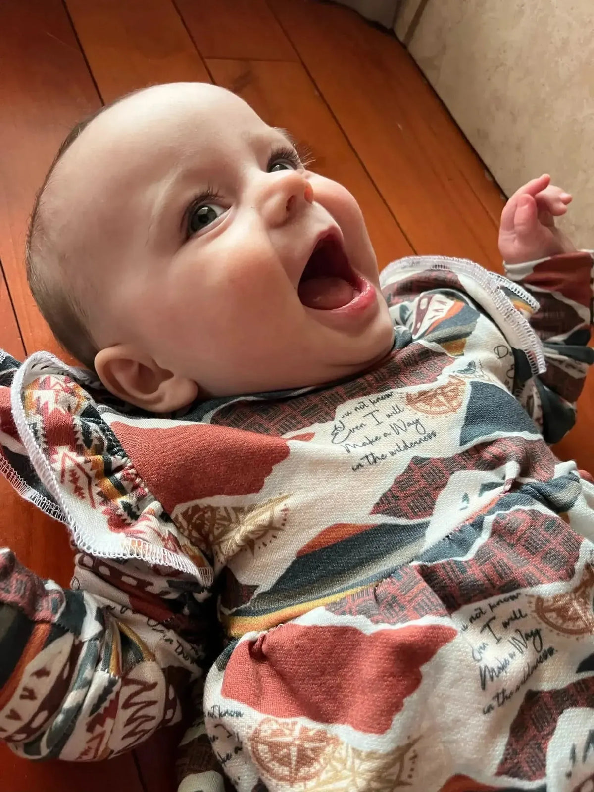 A baby in a patterned "Make a Way" outfit lies on a wooden floor.