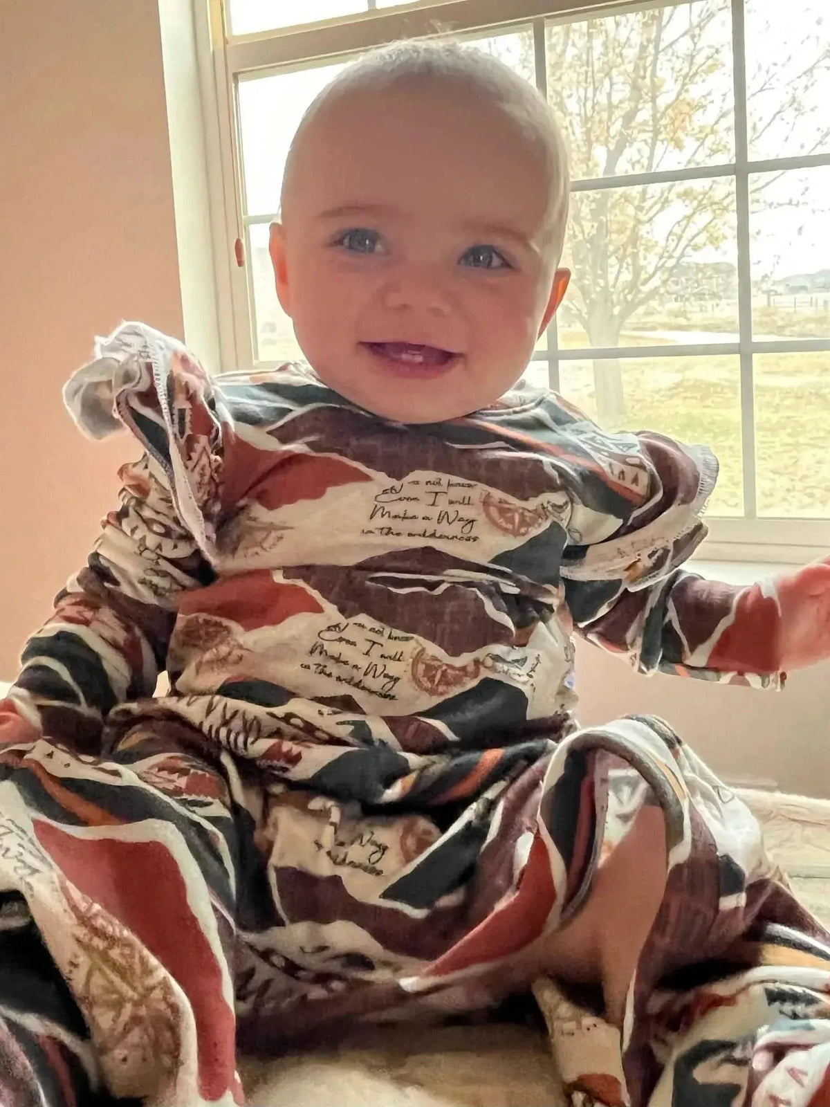 A baby in a "Make a Way" patterned outfit smiles at the camera.