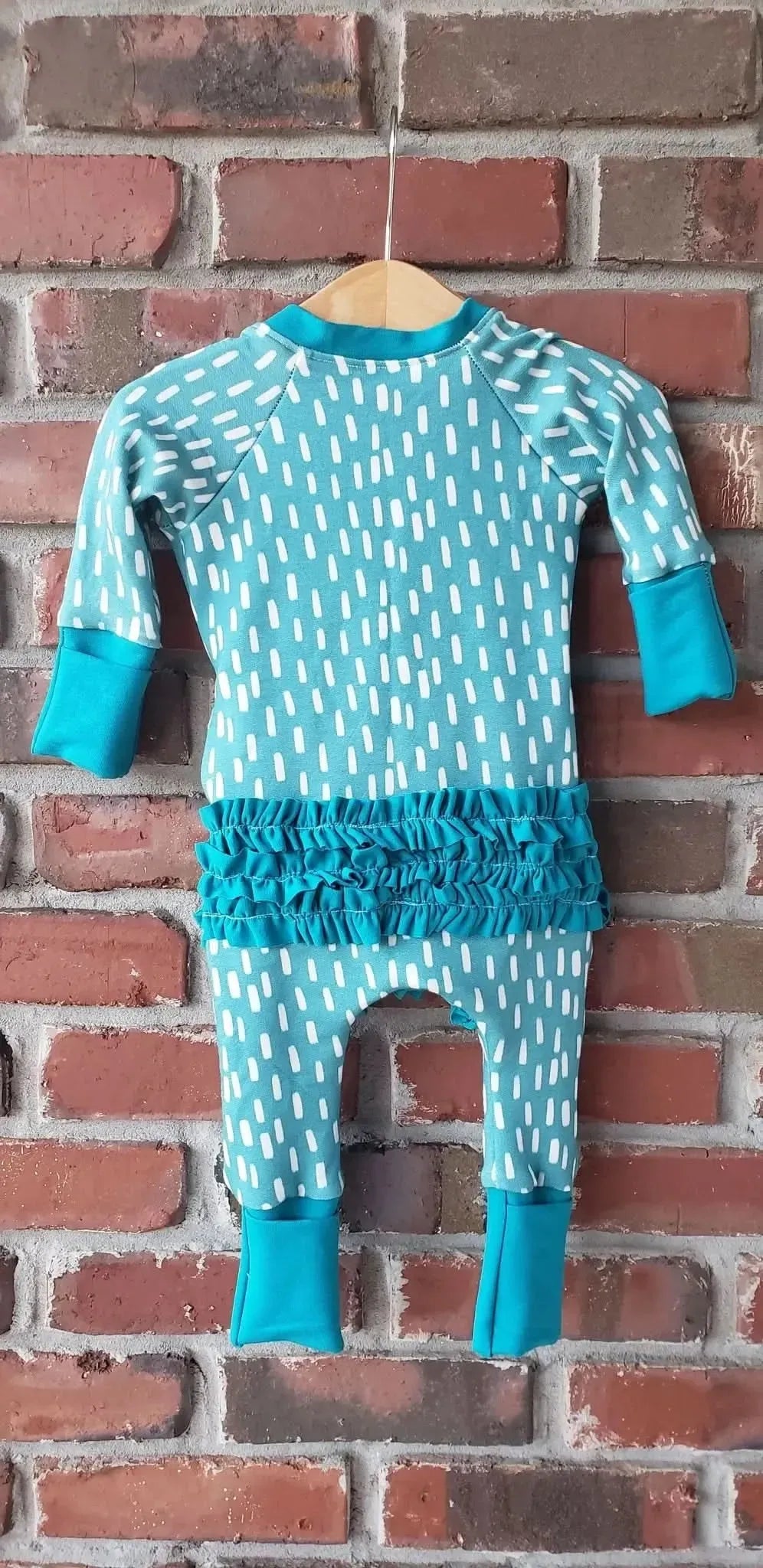 A blue and white patterned baby romper with ruffles hangs on a wooden hanger.
