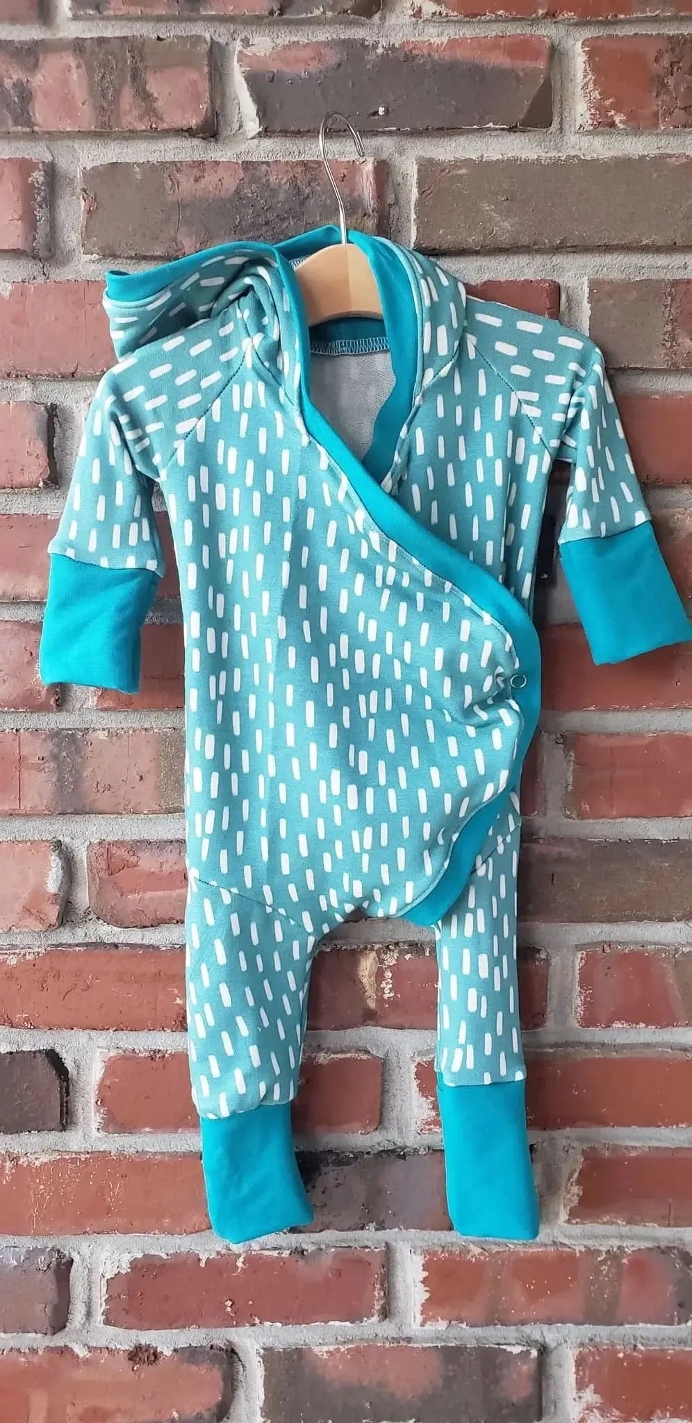 A blue and white patterned baby romper hangs on a wooden hanger against a brick wall.