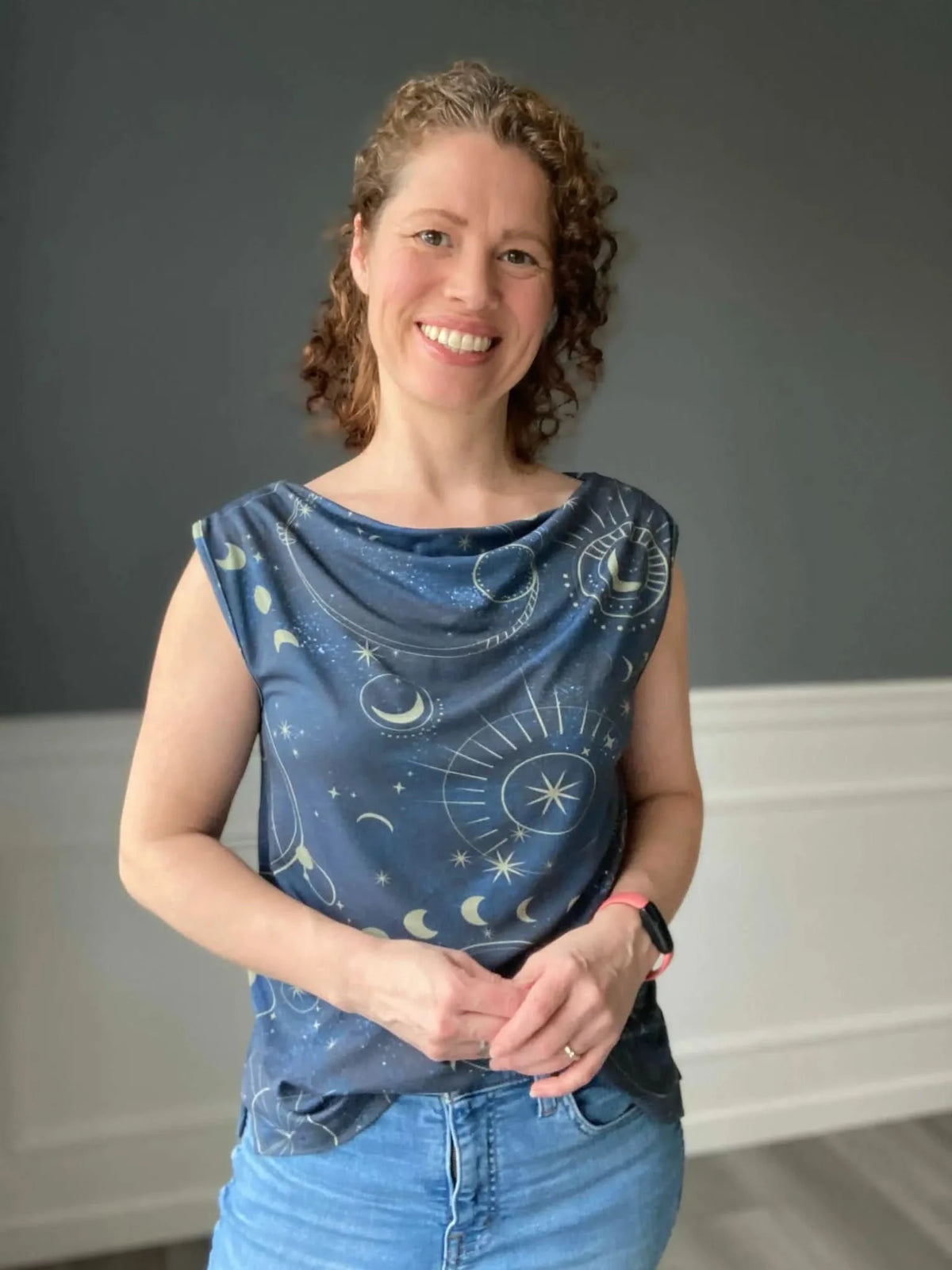 A woman smiles, wearing a navy top with a celestial sky pattern and blue jeans.