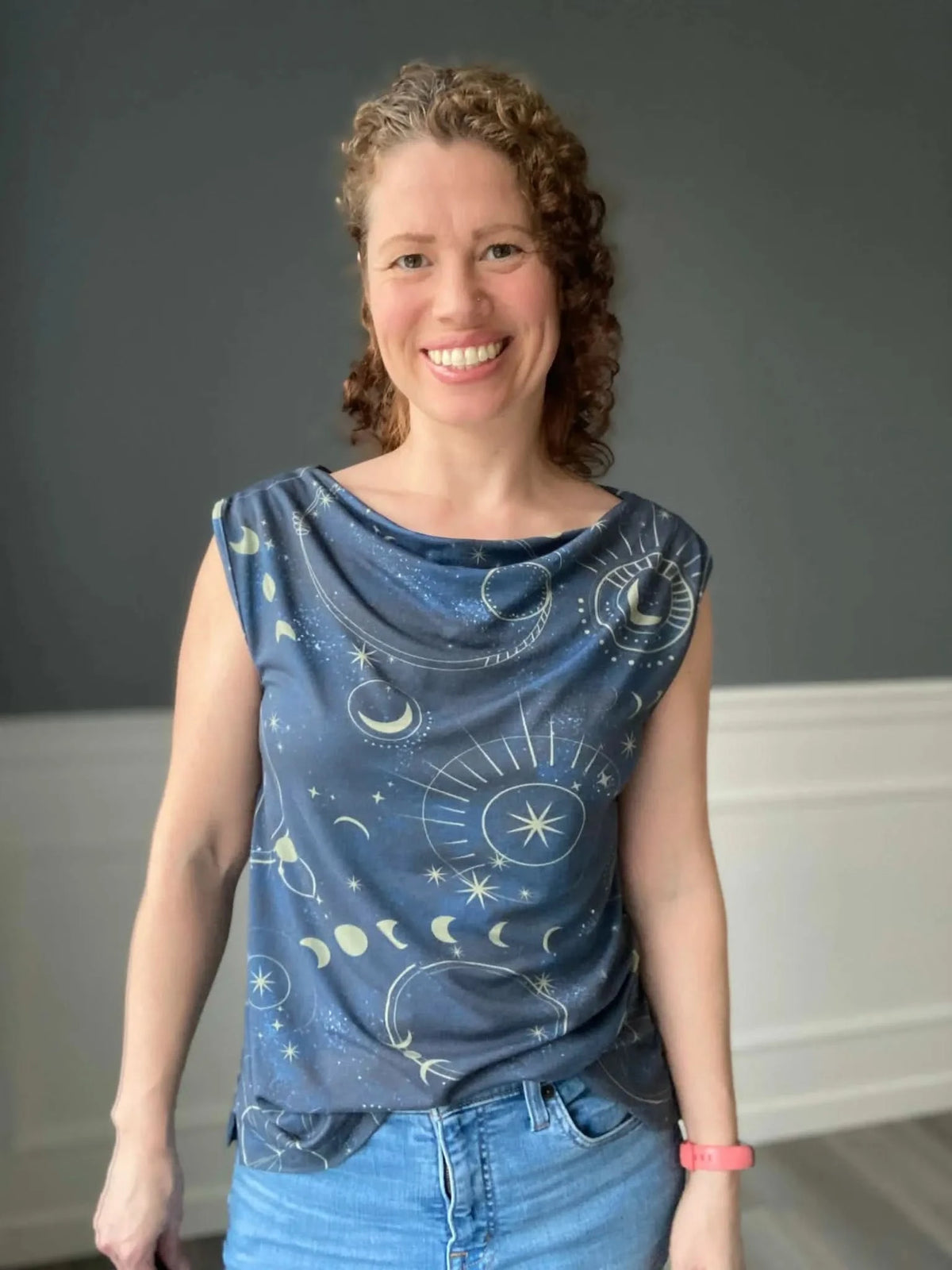 A woman smiles wearing a navy top with a celestial sky pattern and blue jeans.