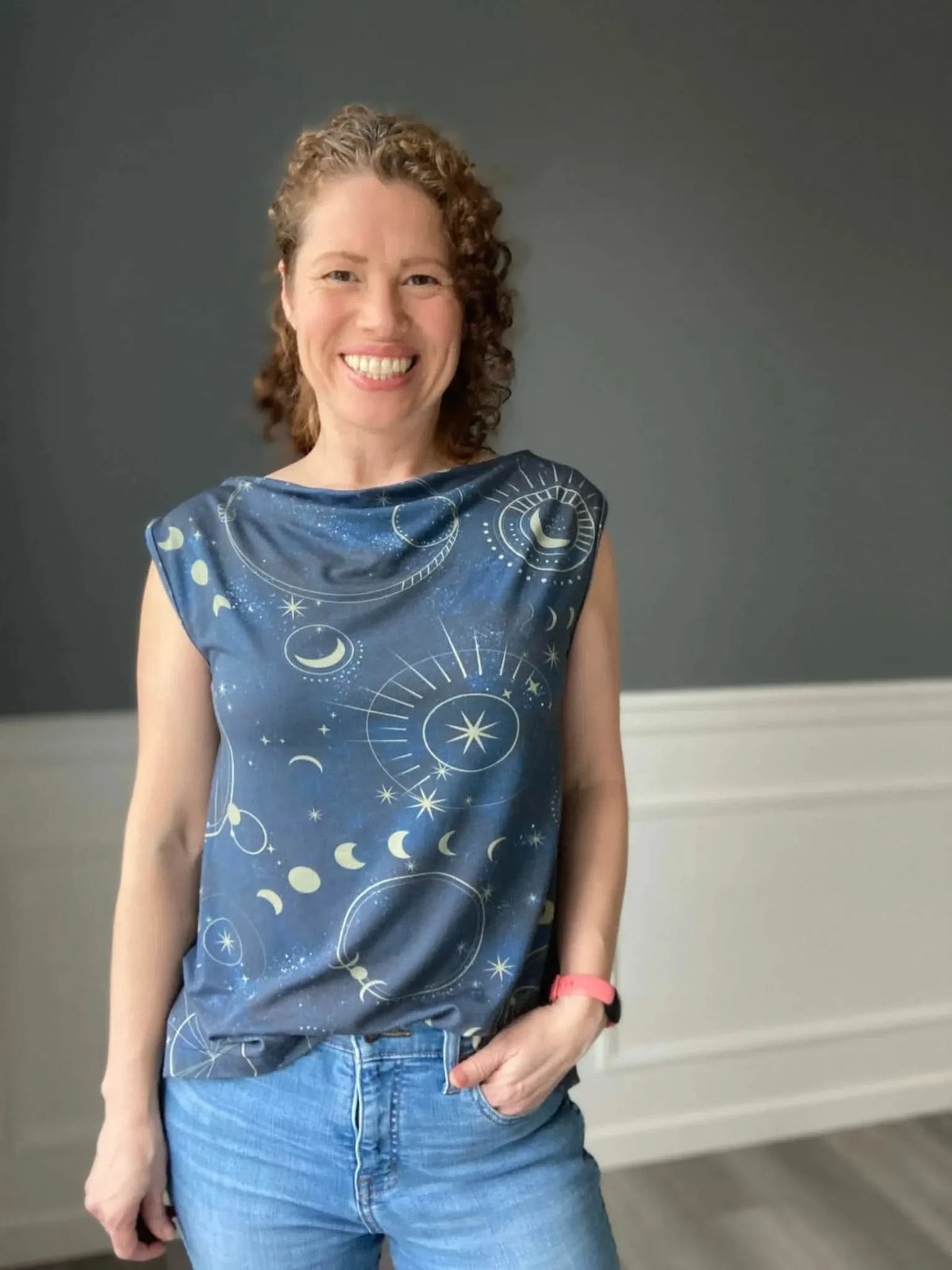 A woman smiles, wearing a navy top with a celestial sky pattern and blue jeans.