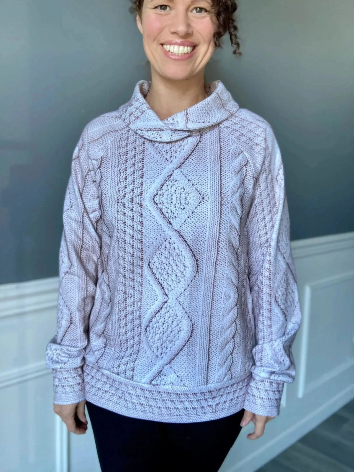 A woman smiles wearing a cream cable knit sweater with a cowl neck.