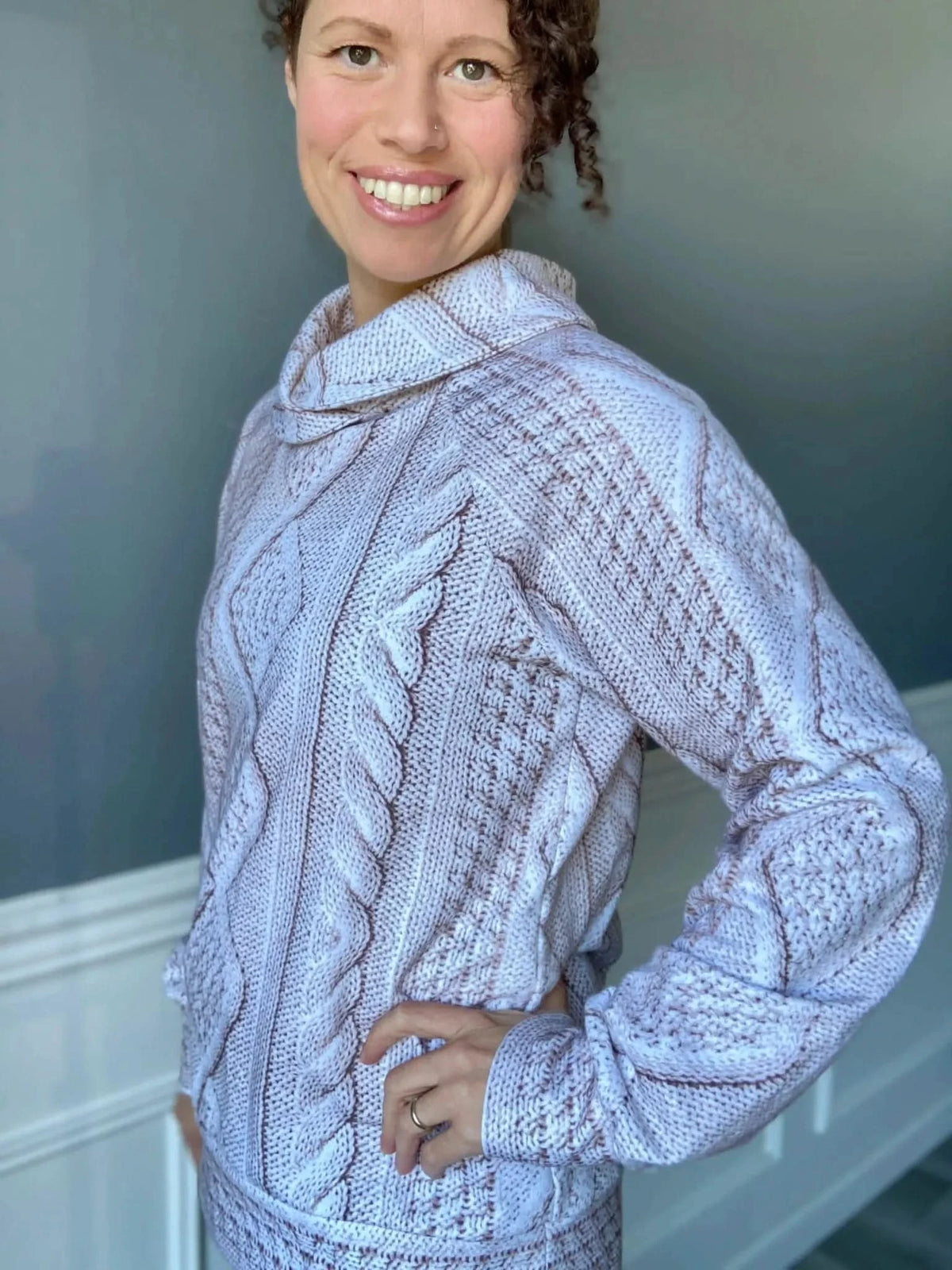 A woman smiles, wearing a cream cable knit sweater with brown accents.