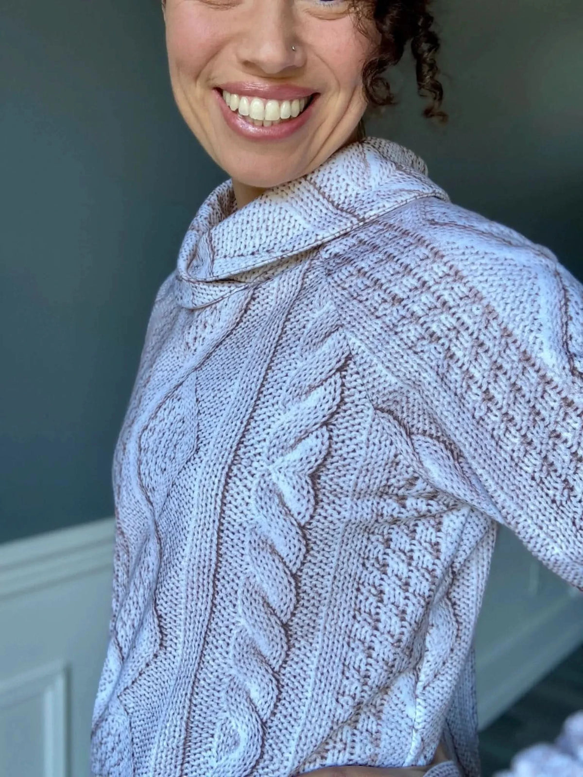 A woman smiles, wearing a cream cable knit sweater with a cowl neck.