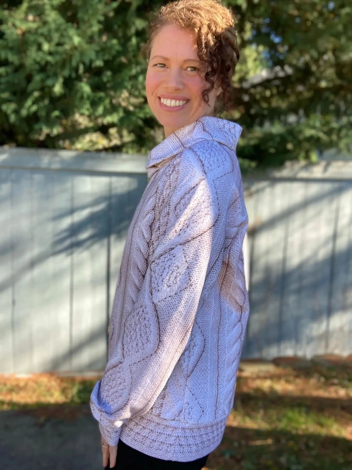 A woman smiles, wearing a cream cable knit sweater with a collared neckline.