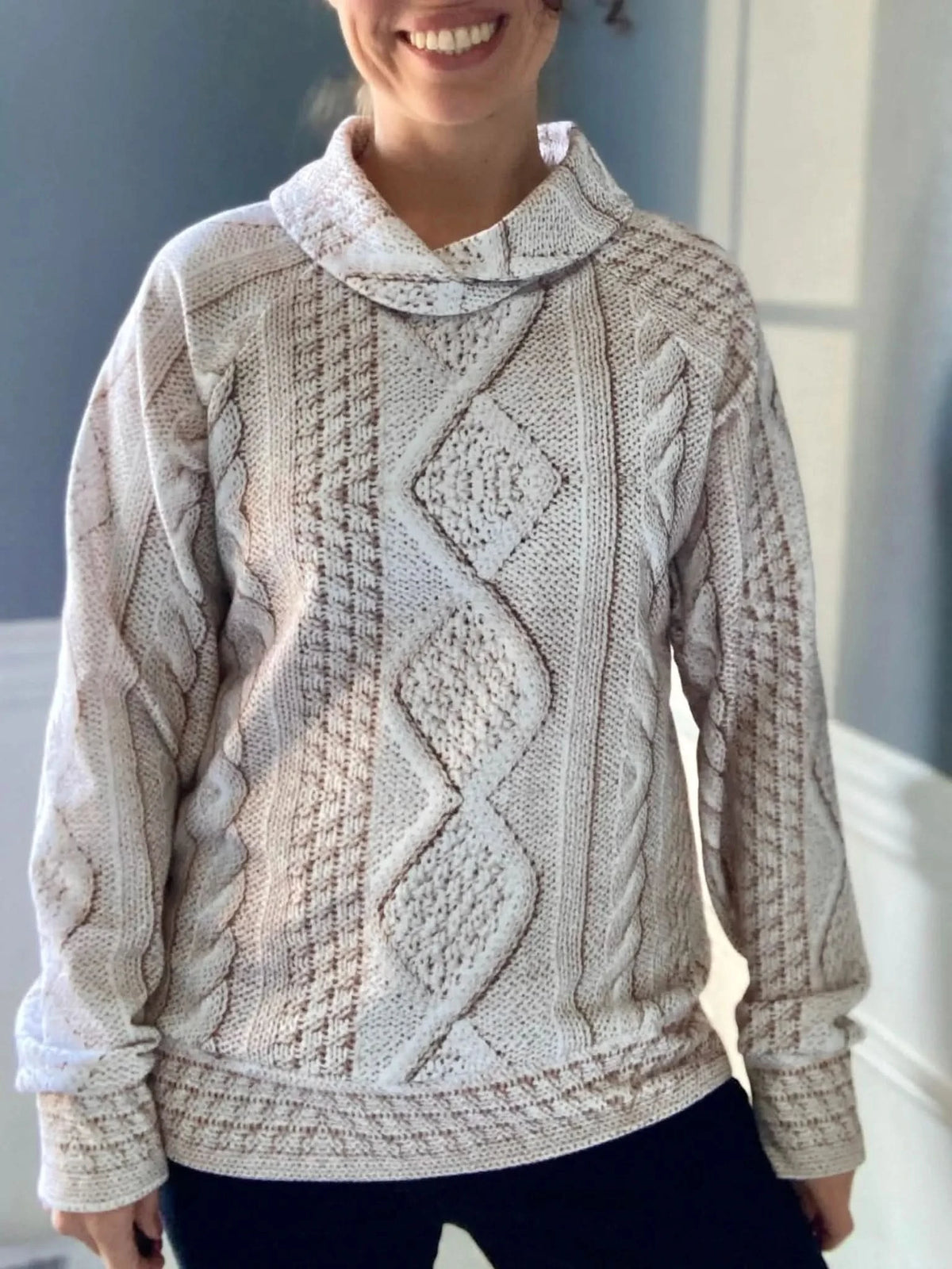 A person wears a cream cable knit sweater with a cowl neck.