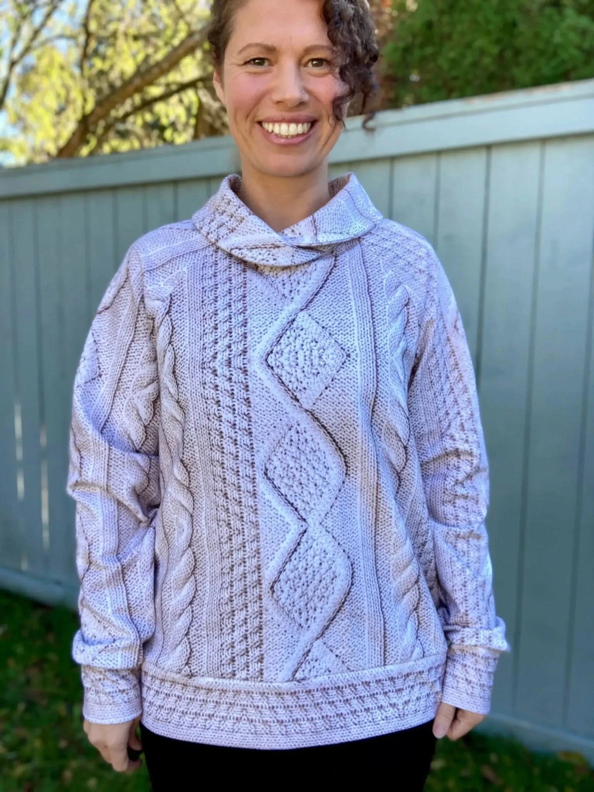 A woman smiles wearing a cream cable knit sweater with a cowl neck.