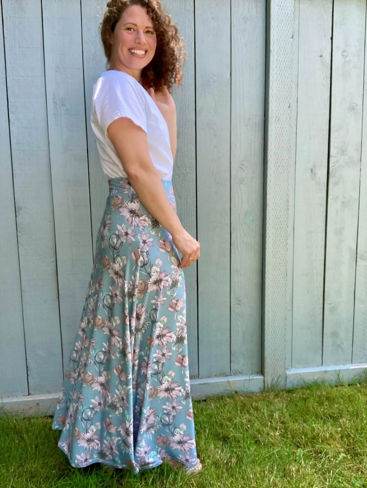 A woman in a white shirt and a floral maxi skirt smiles in front of a fence.