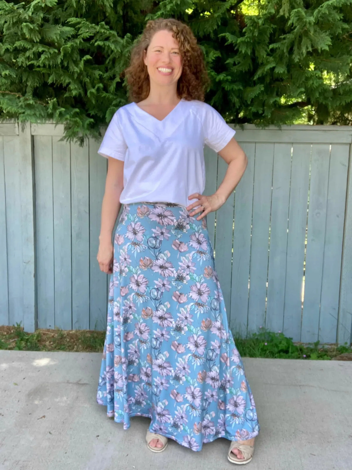 A woman smiles, wearing a white top and a blue floral maxi skirt.