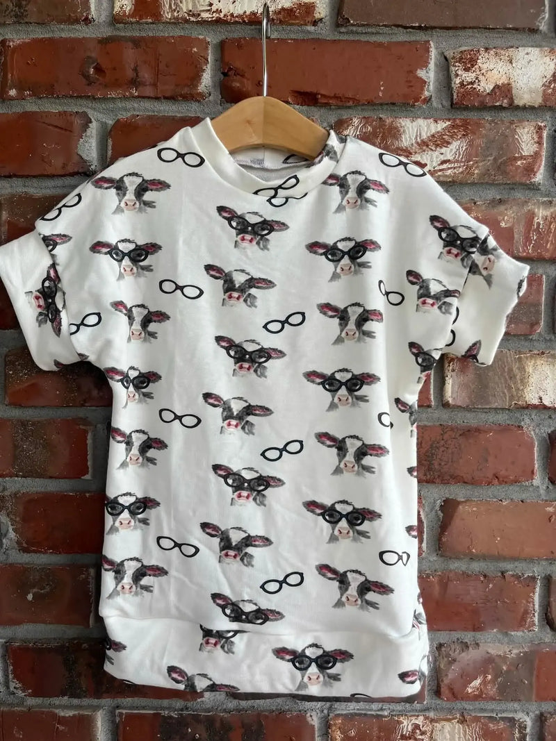 A white t-shirt with a pattern of cows, some wearing glasses, hangs on a hanger.