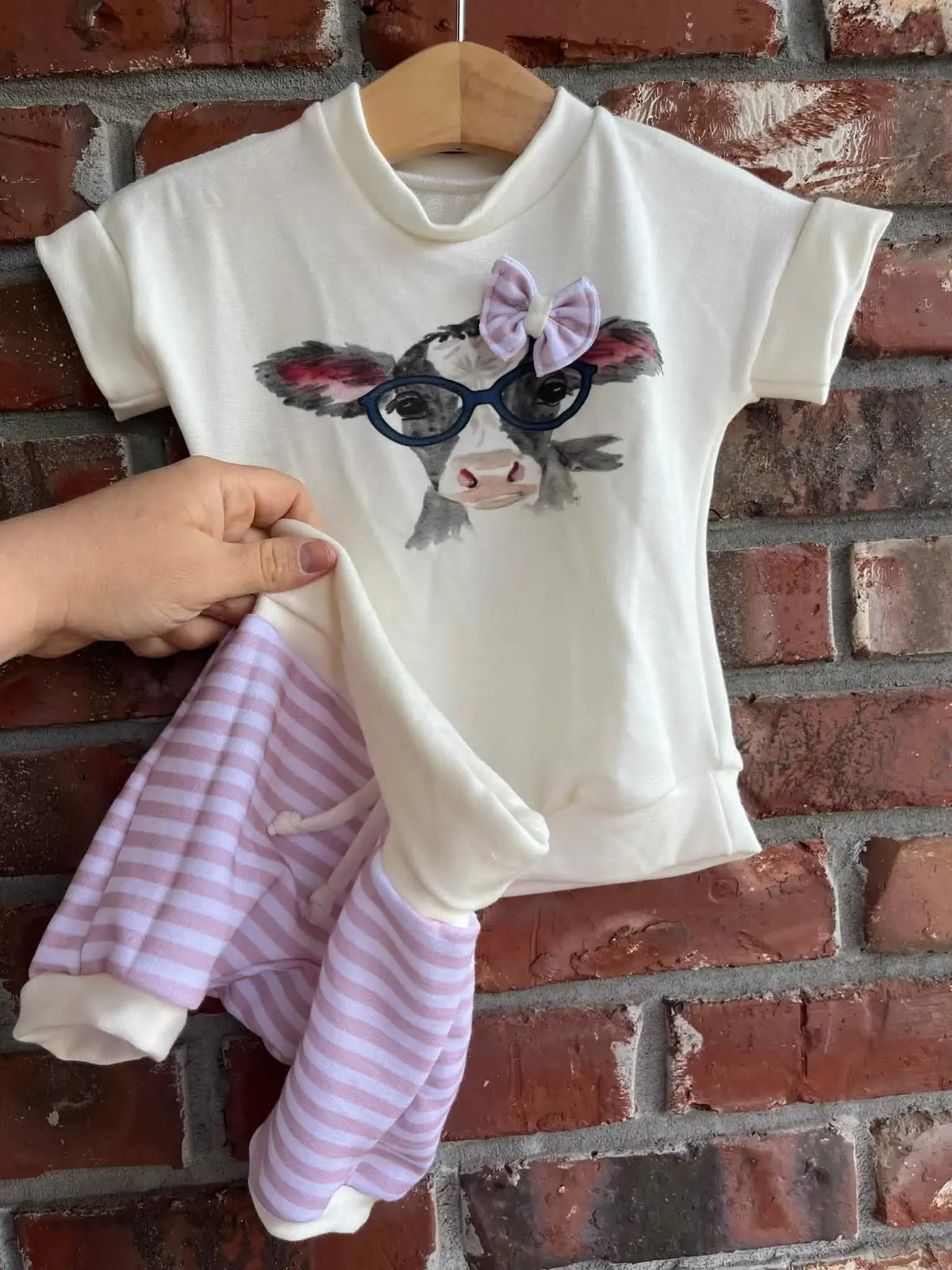 A white top with a cow wearing glasses and a pink bow, with striped pants.