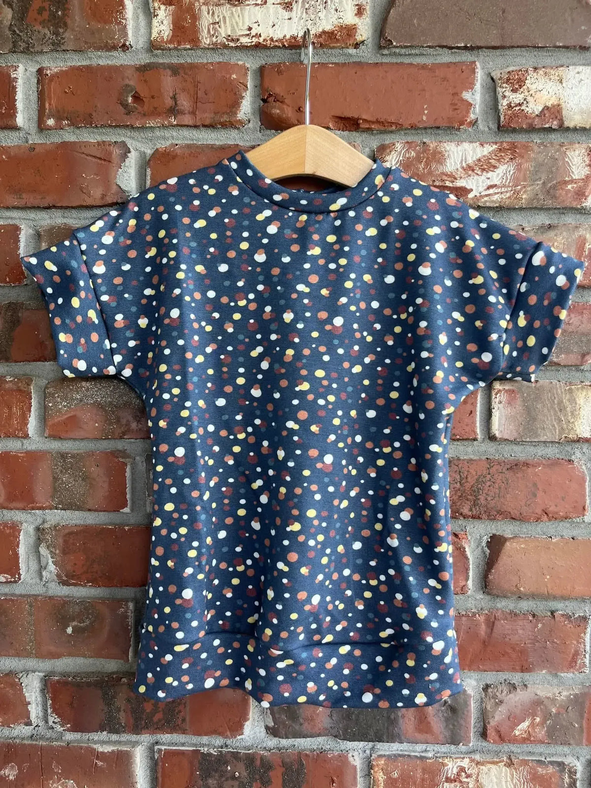 A navy shirt with colorful sprinkles hangs on a wooden hanger against a brick wall.