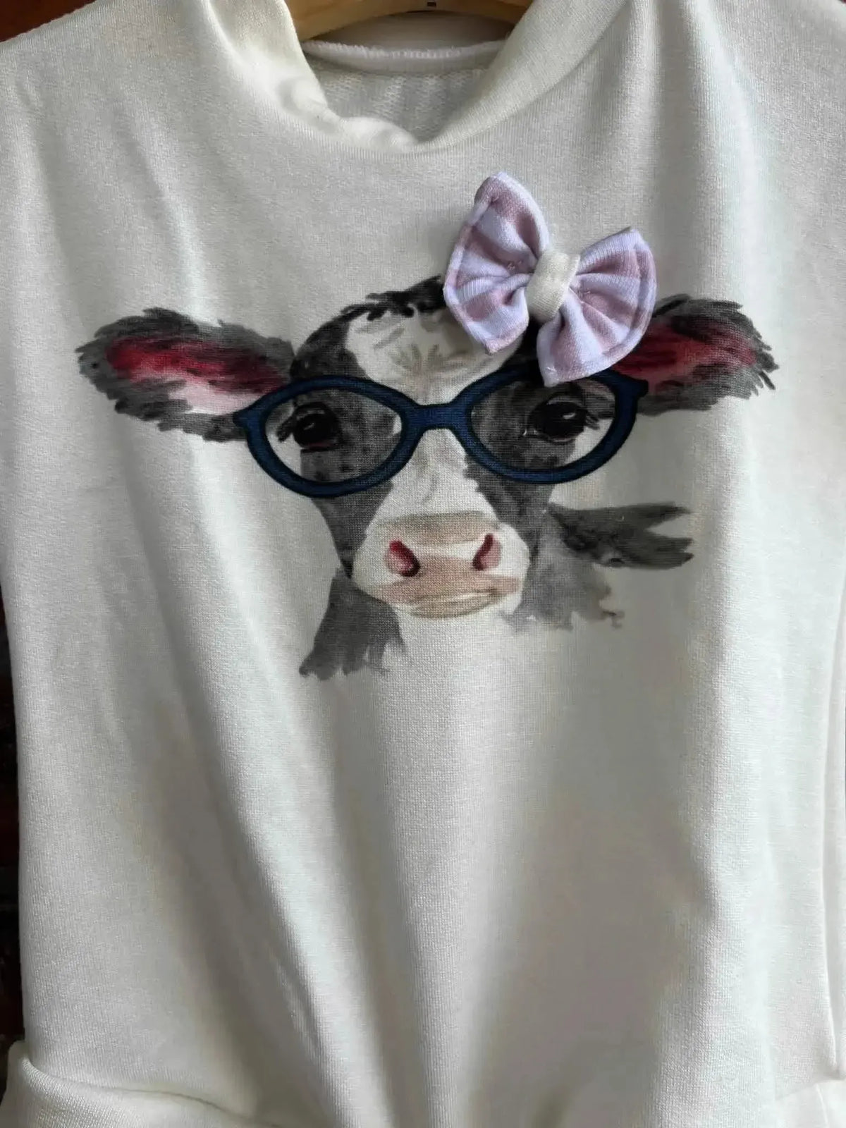 White fabric panel with a cute black-and-white cow face wearing glasses and a purple bow.