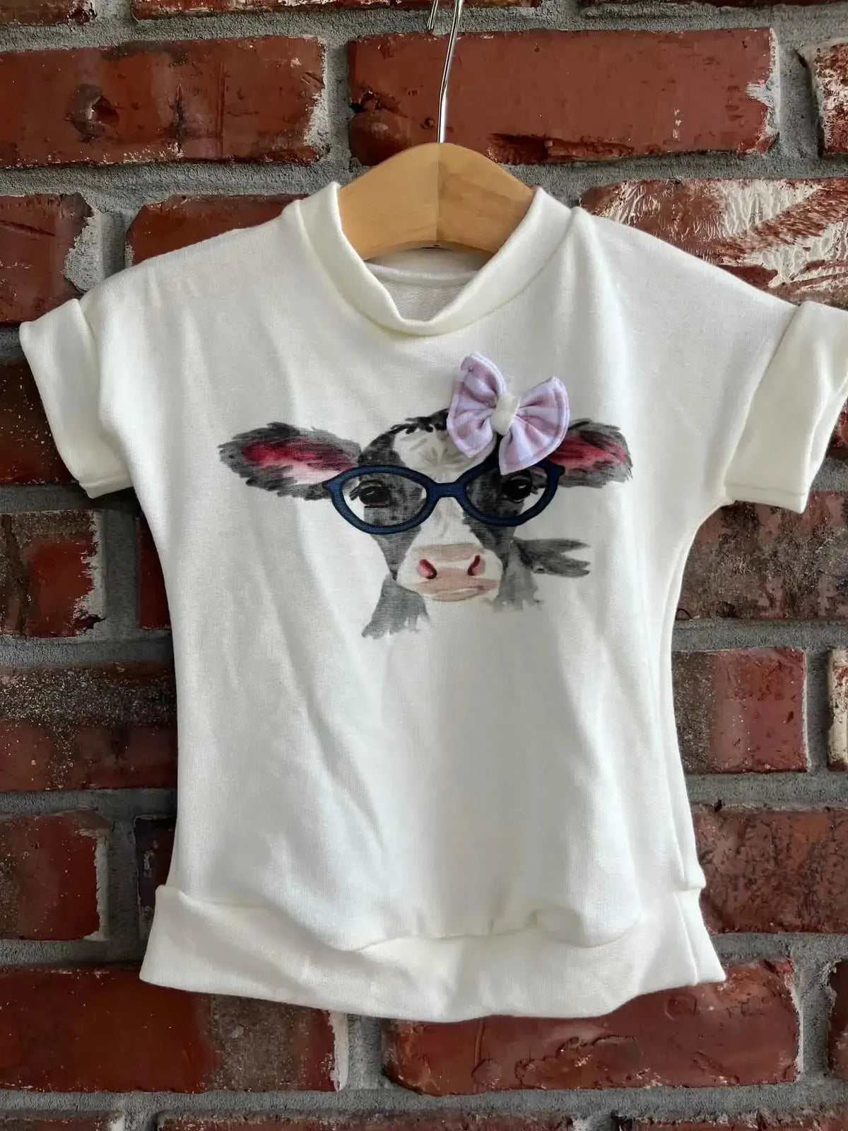 White short-sleeve baby shirt with a printed cow face wearing red glasses and a purple bow.
