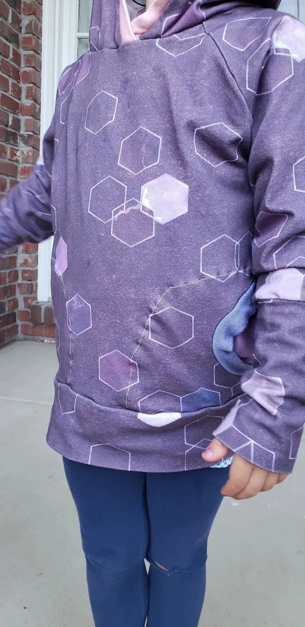A child wears a purple hoodie with a geometric pattern and blue pants.
