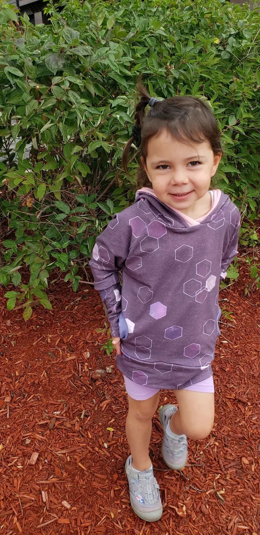 A young girl wears a burgundy hoodie with geometric patterns and light purple shorts.