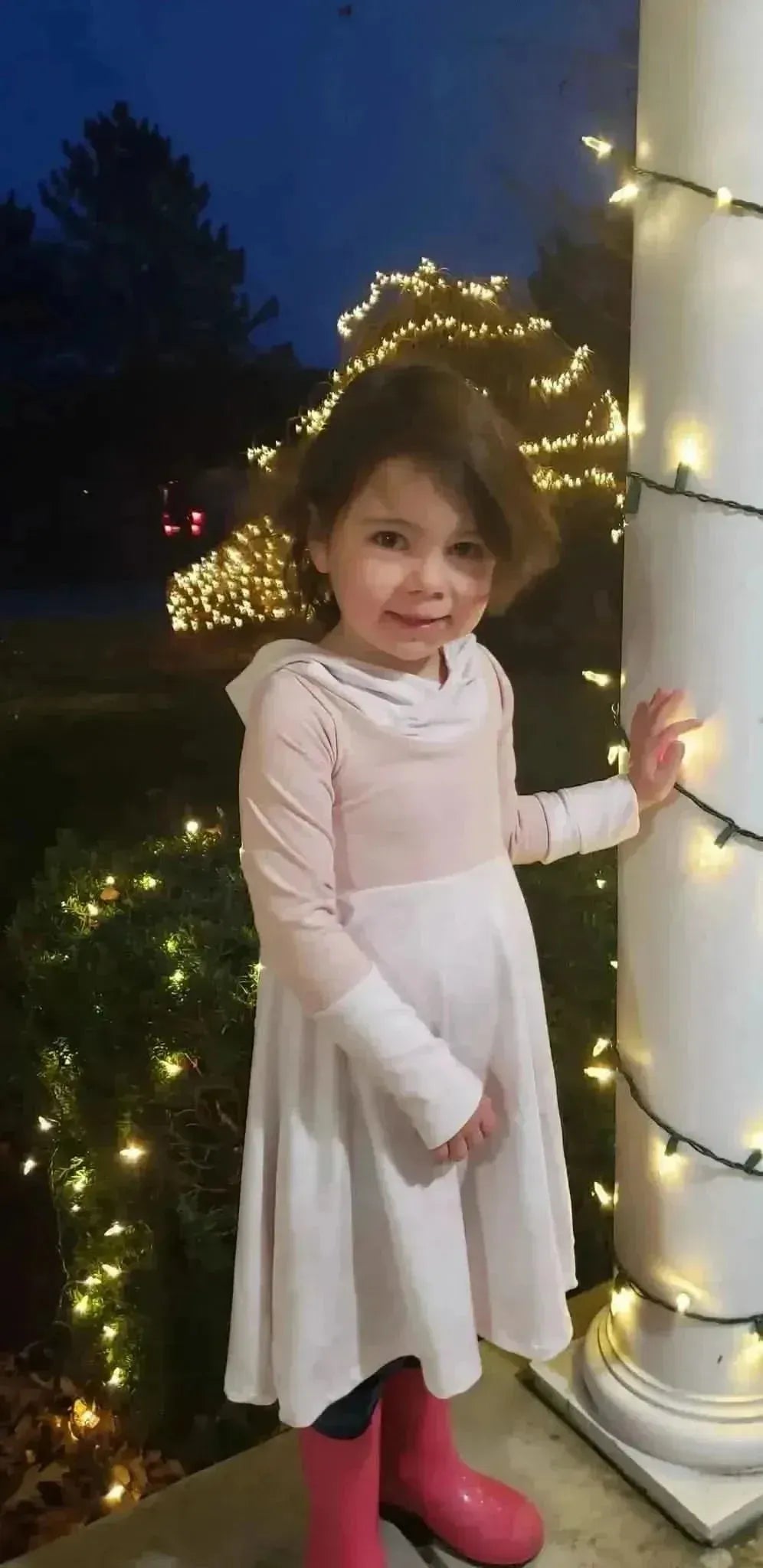 A young girl in a pink and white dress and pink boots stands by a lit pillar.