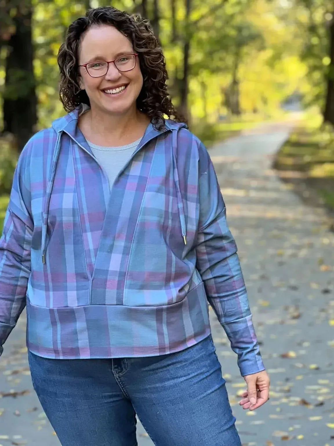 A woman in a dark Christmas plaid hoodie and jeans smiles on a path with autumn leaves.