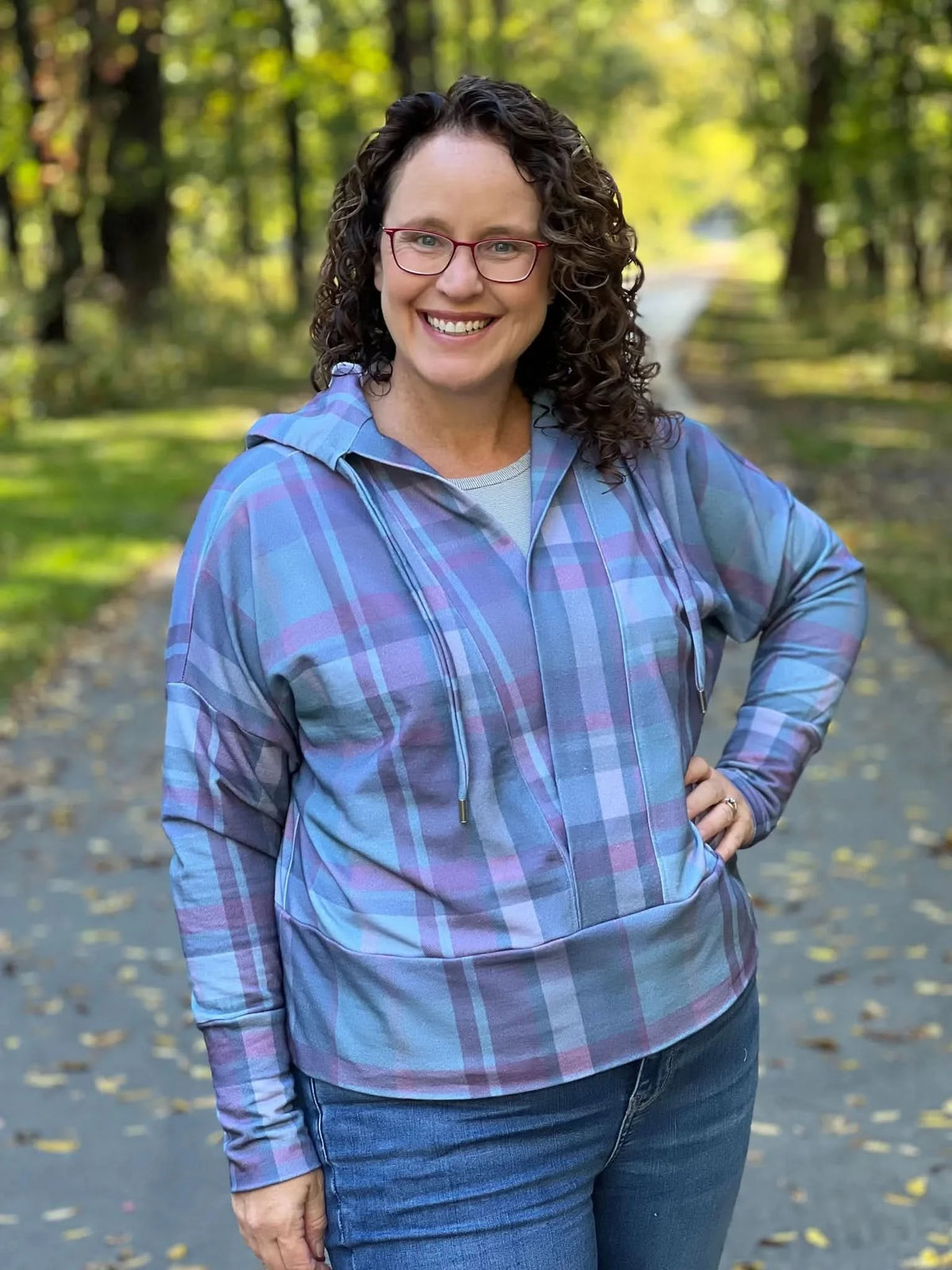 Woman in a blue and purple plaid hoodie and jeans, smiling on a path.
