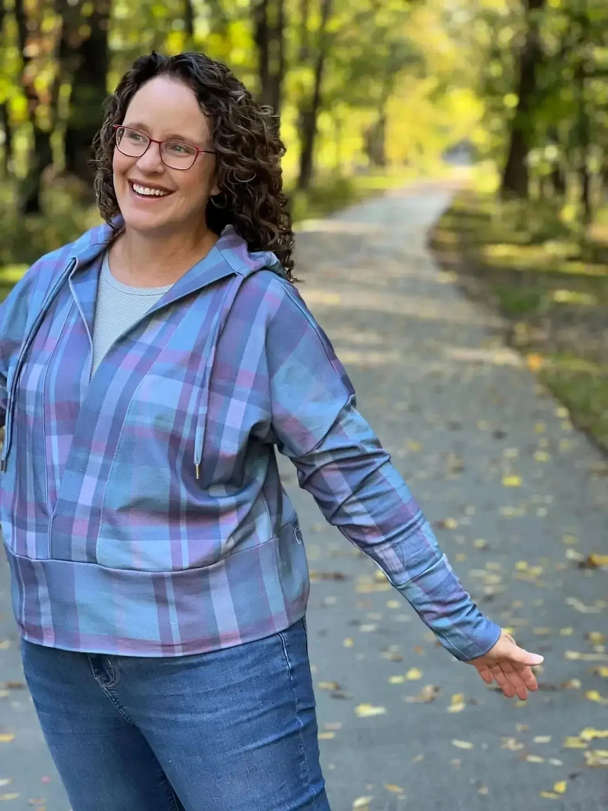 A woman in a blue and purple plaid hoodie smiles on a path with autumn leaves.