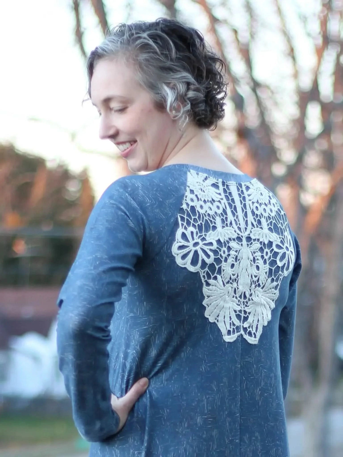 A woman in a blue top with white lace on the back smiles, looking over her shoulder.