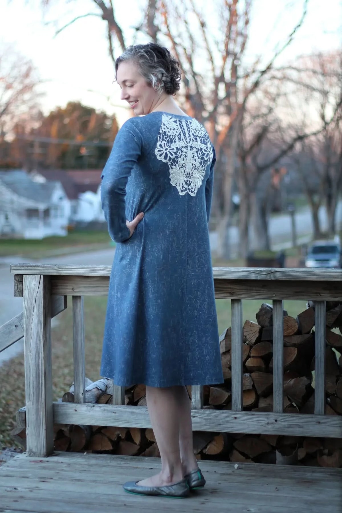 A woman in a blue dress with white lace on the back stands on a wooden porch.