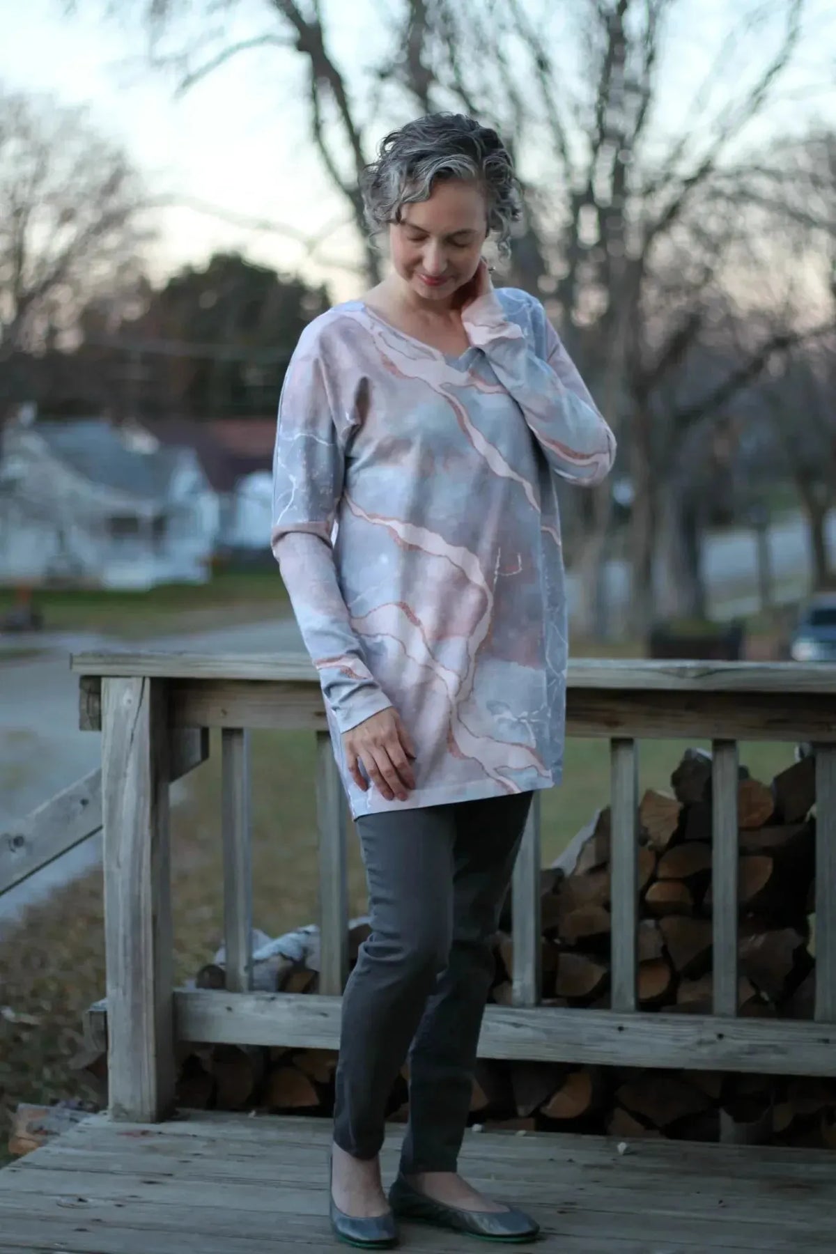 Woman wearing a long-sleeved pink marble print tunic with gray pants on a porch.