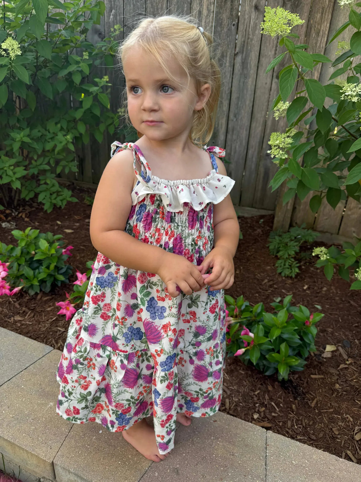A young girl in a floral dress stands barefoot on a stone patio.