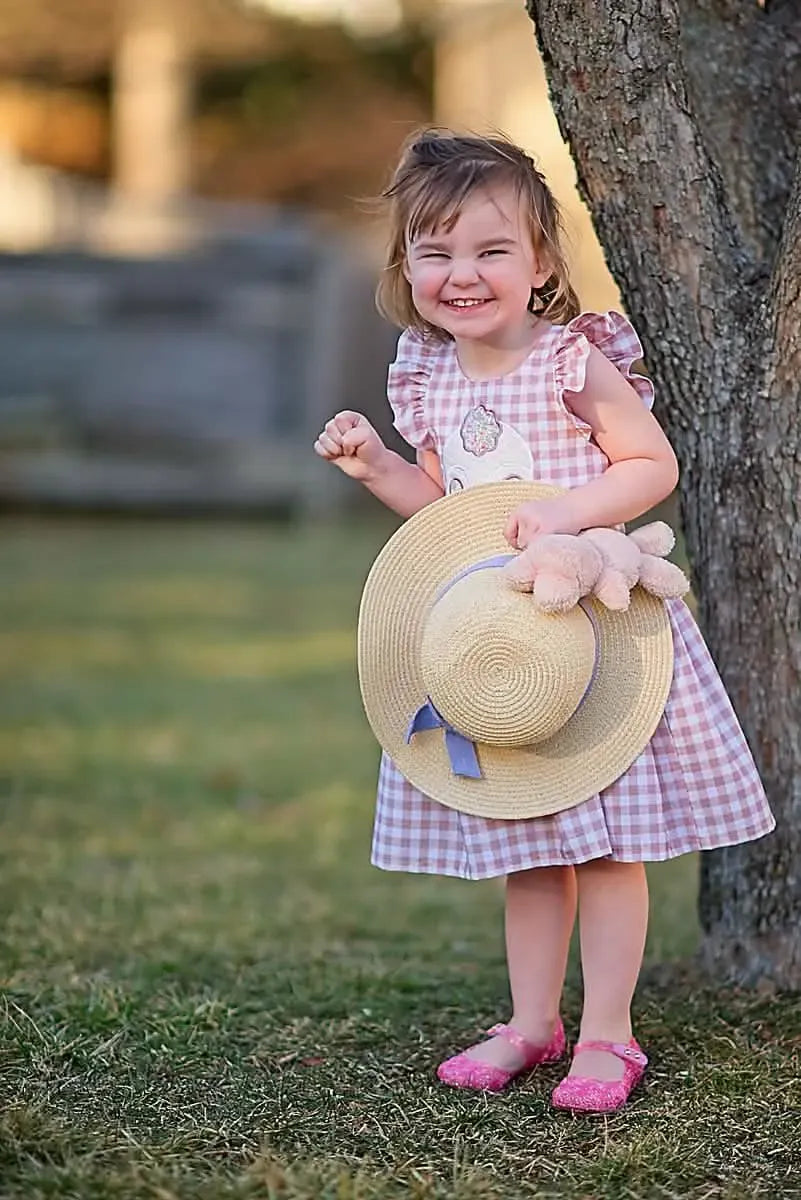 A young girl in a tan gingham dress holds a straw hat with a blue ribbon.