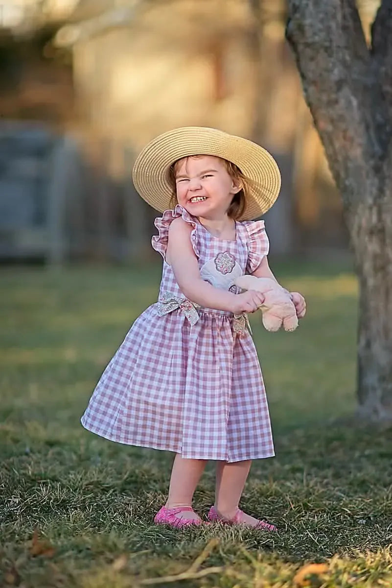 A young girl in a tan gingham dress and straw hat smiles while holding a toy.