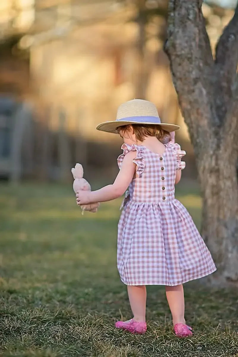 A child in a tan gingham dress and straw hat holds a stuffed bunny.