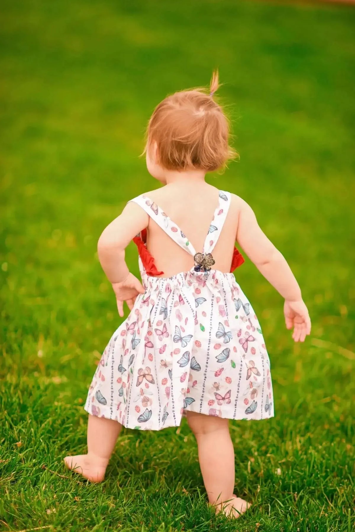 A toddler in a white dress with colorful bug and butterfly patterns stands on green grass.