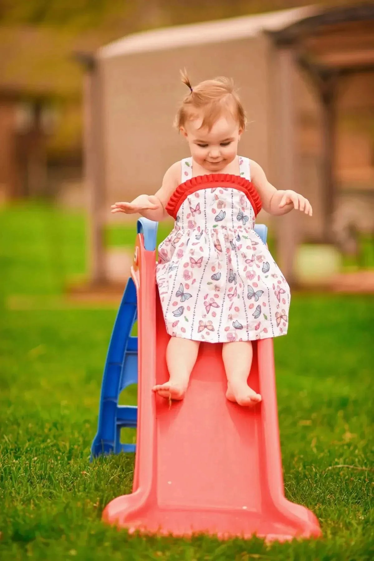 Toddler in a white dress with colorful bug and butterfly patterns on a red slide.