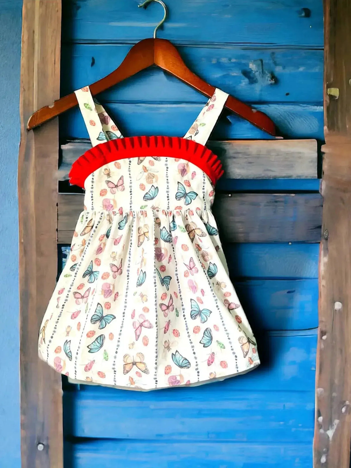 White dress with red ruffle straps and butterfly print, hanging on a wooden hanger against a blue wall.