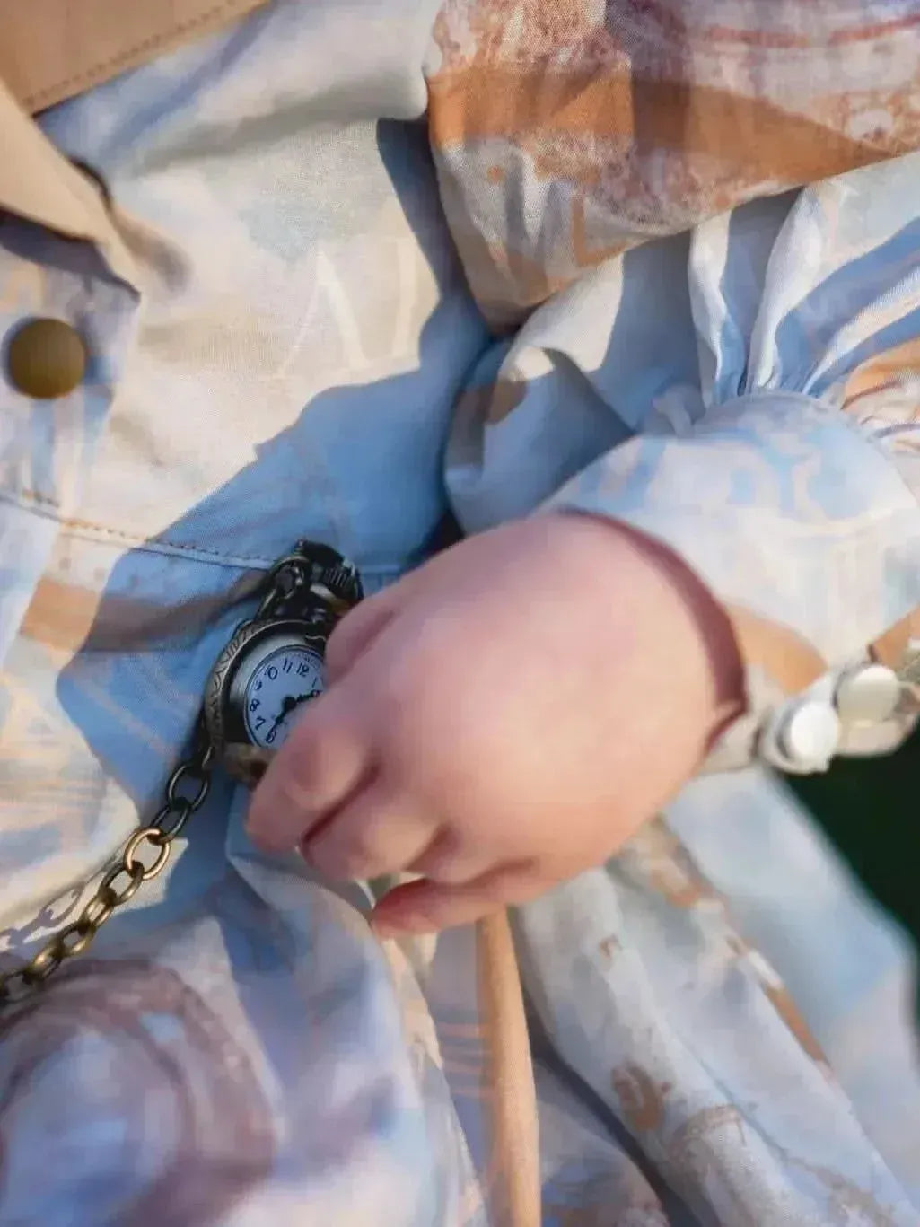 A baby's hand holds a pocket watch against a light blue and brown patterned fabric.