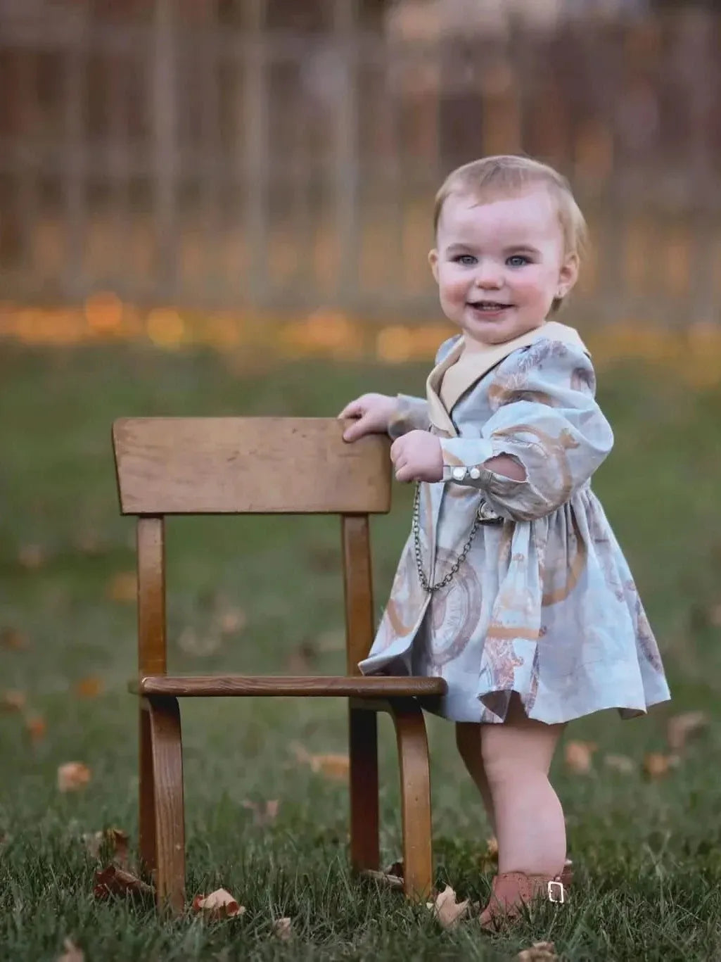 A baby in a patterned dress stands next to a small wooden chair outdoors.