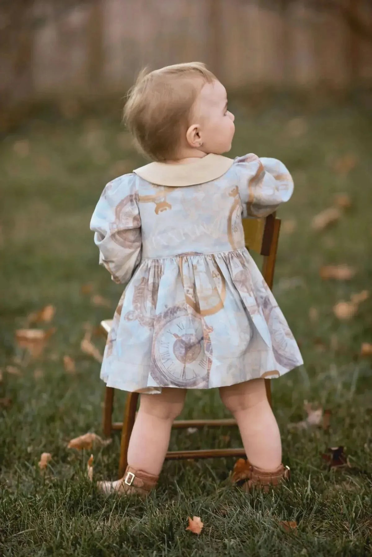 A baby in a light blue dress with gold clock patterns stands with her back to the camera.