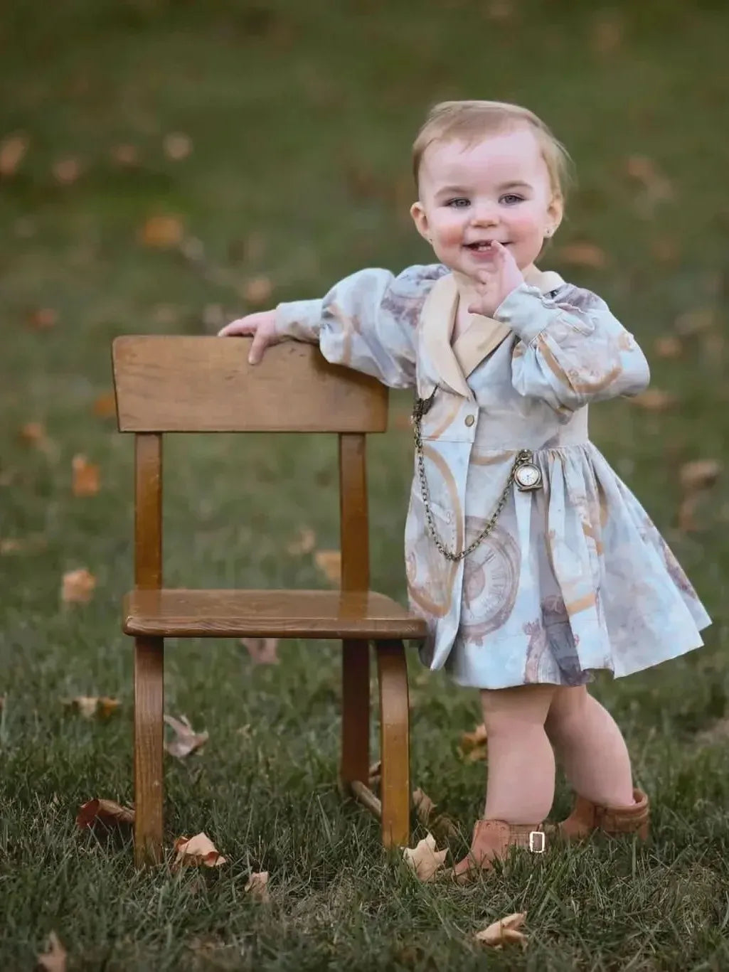 A baby in a light blue dress with a clock pattern leans on a small wooden chair.