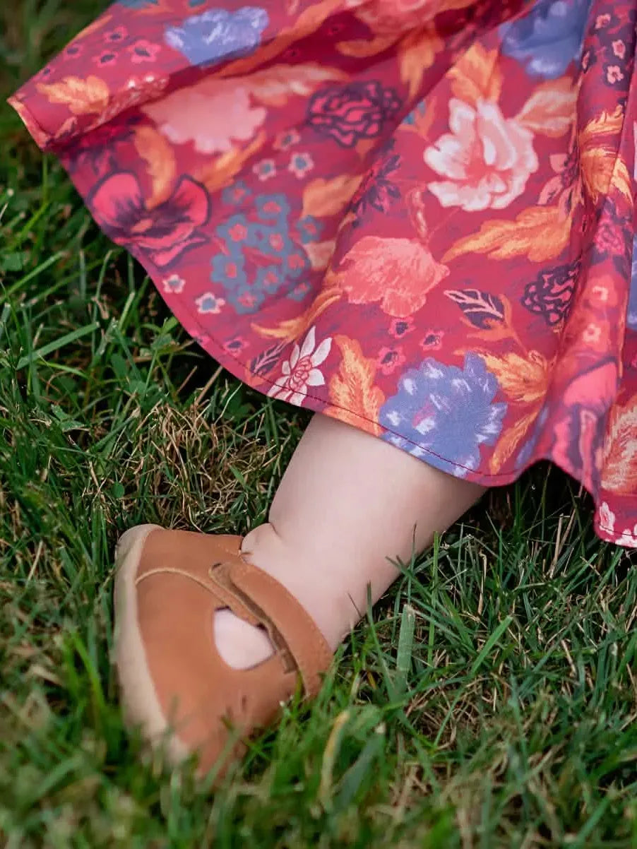 A child's leg and foot in a brown shoe, with a red floral dress on green grass.