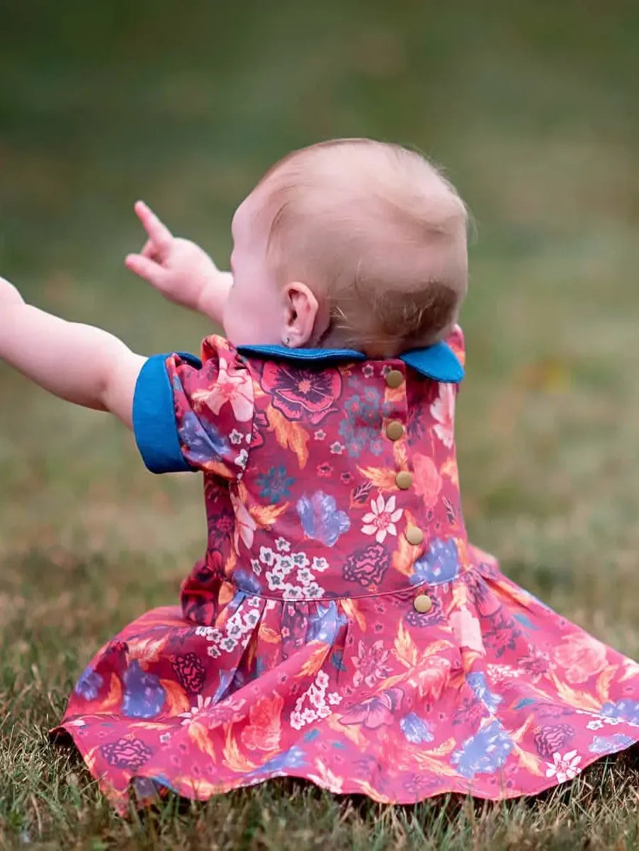 A baby in a red floral dress with blue trim sits on grass, pointing to the left.