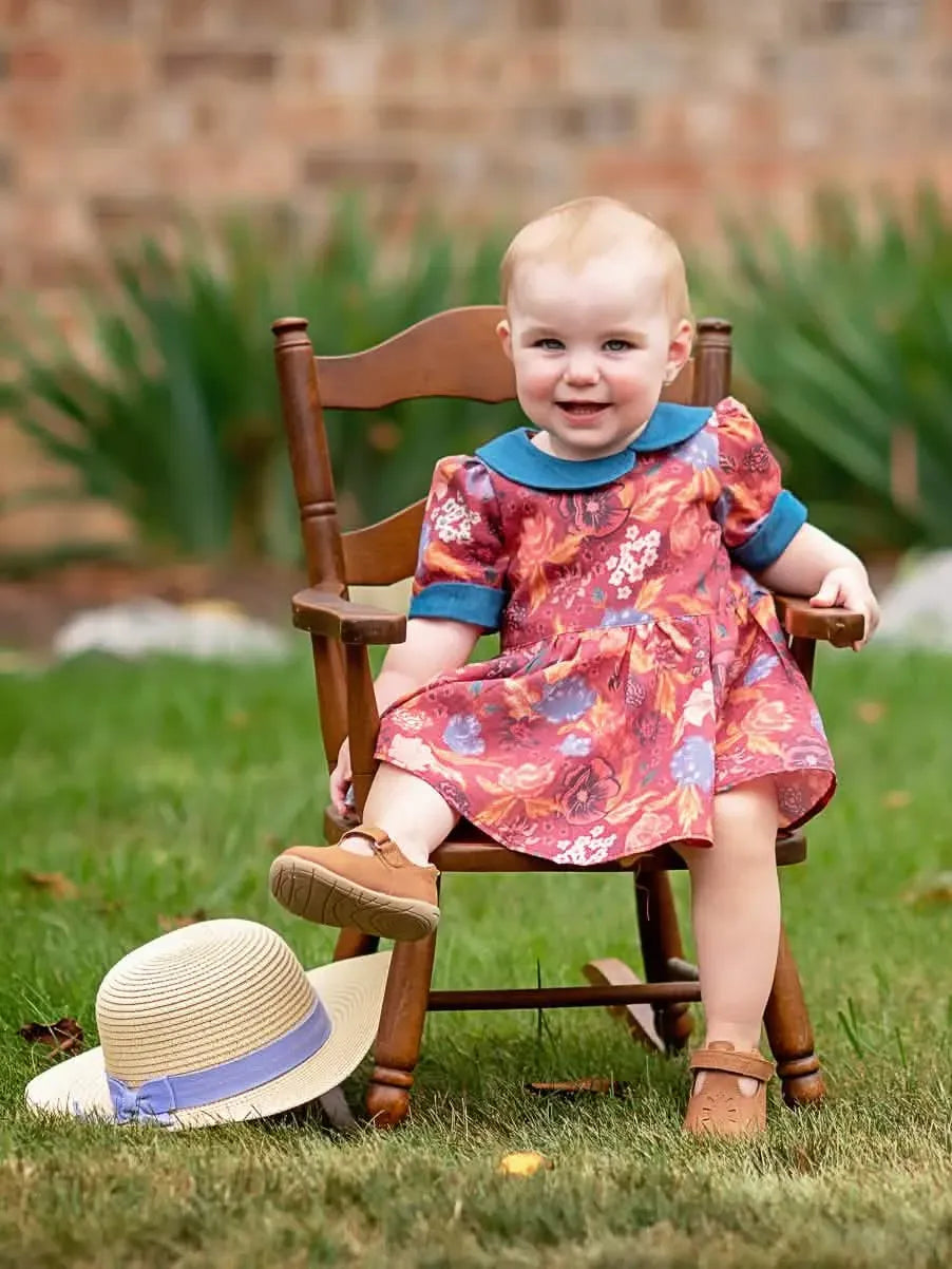 A baby in a floral dress with a blue collar sits in a wooden rocking chair.