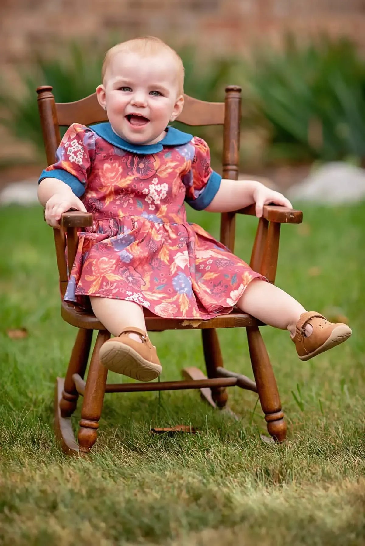 A baby in a floral dress with a blue collar sits in a wooden rocking chair.