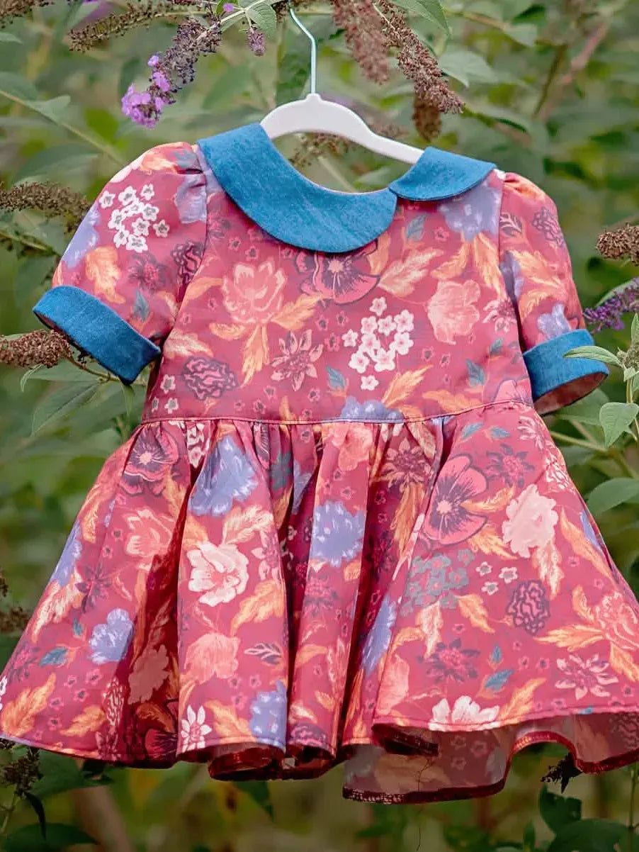 A floral dress with a blue collar and cuffs hangs on a white hanger amidst green foliage.
