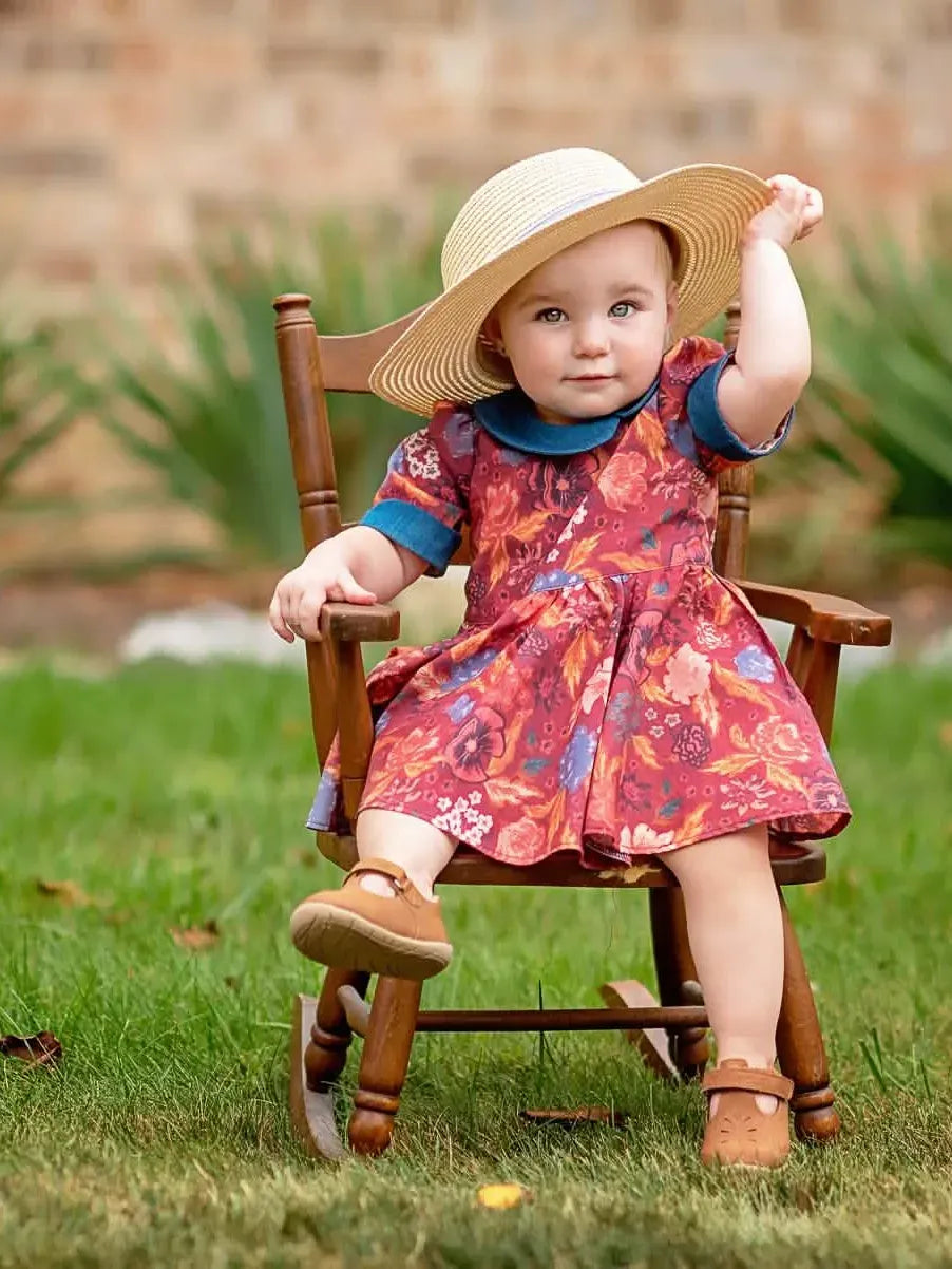 A baby in a floral dress and straw hat sits in a wooden rocking chair on grass.