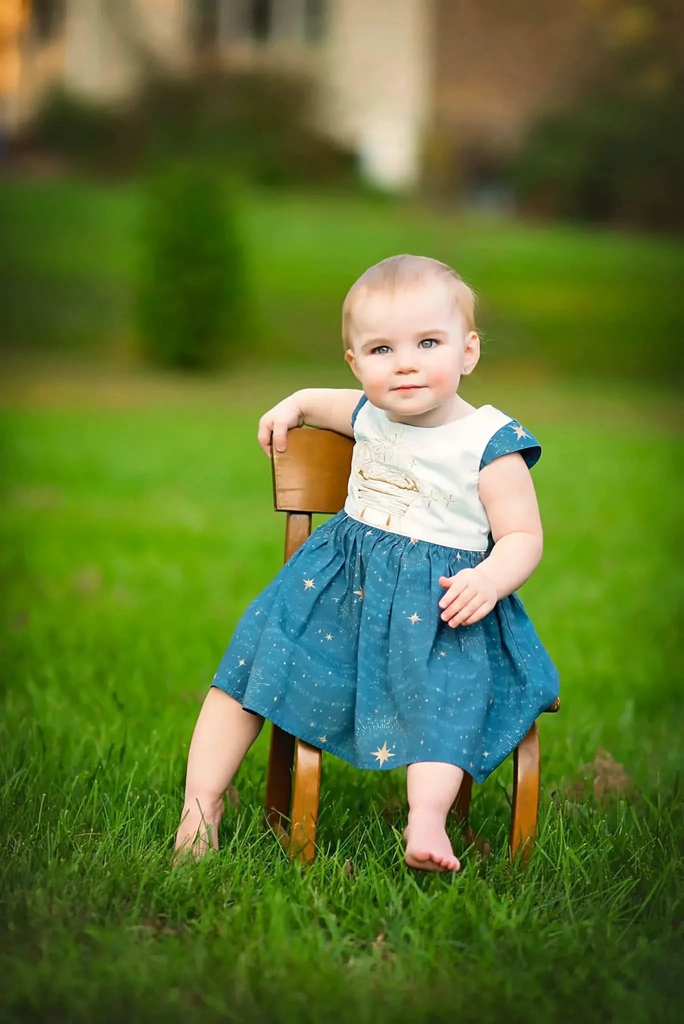 A baby in a blue dress with gold stars sits on a small wooden chair in grass.