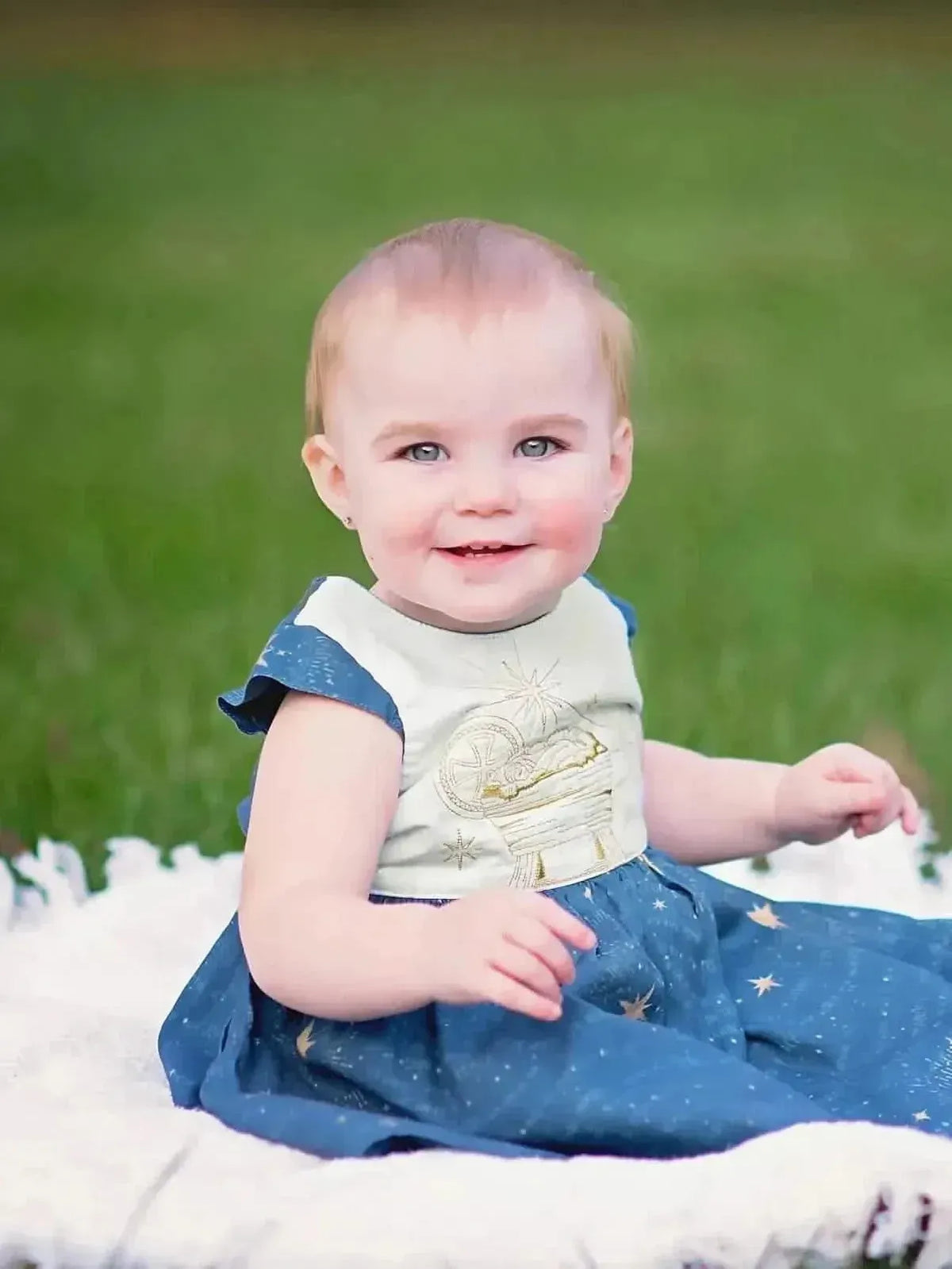 Baby in celestial blue dress with gold moon and stars on white fabric, sitting on grass.