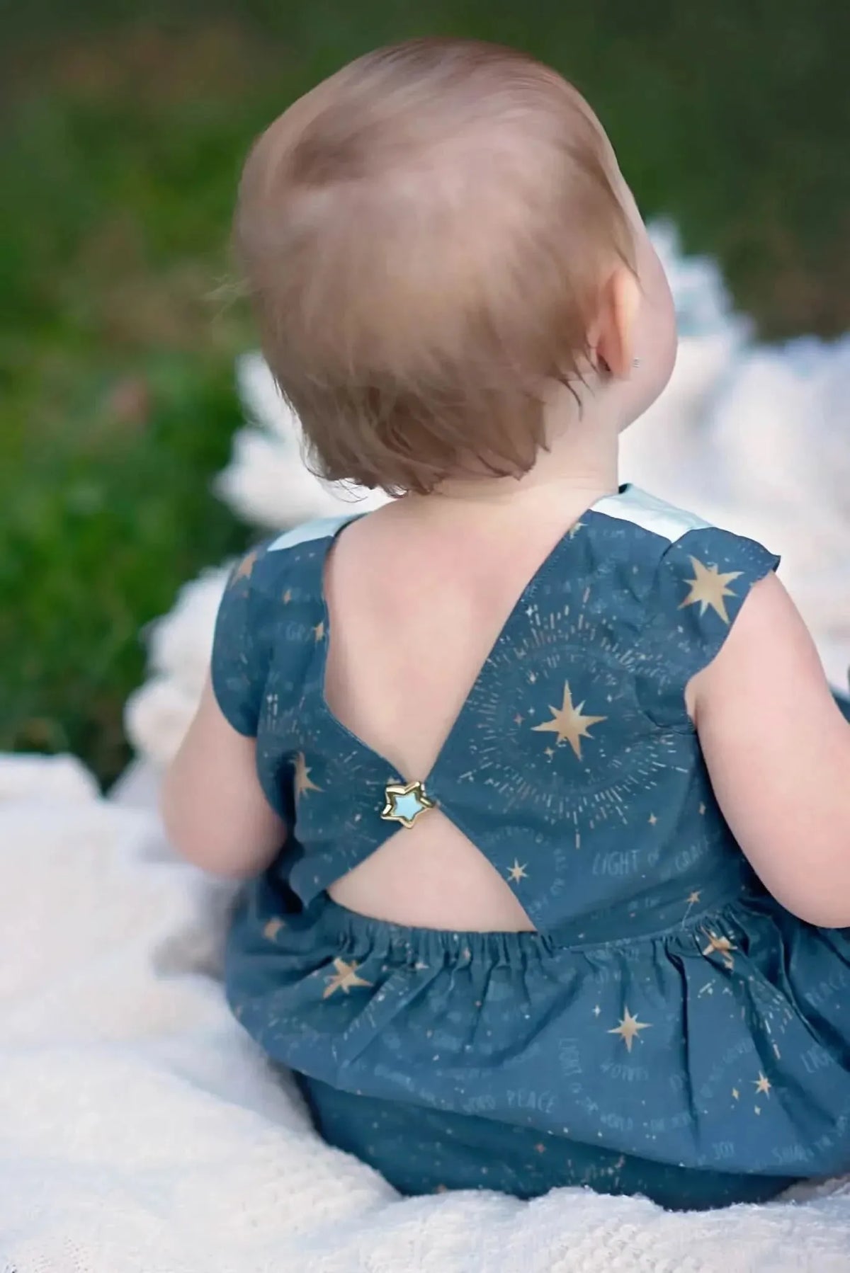 Baby in celestial blue dress with star print on grassy blanket.